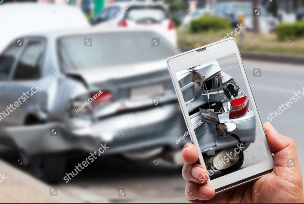 A person holding a smartphone captures a photo of a car with severe rear-end damage after an accident on a city street.